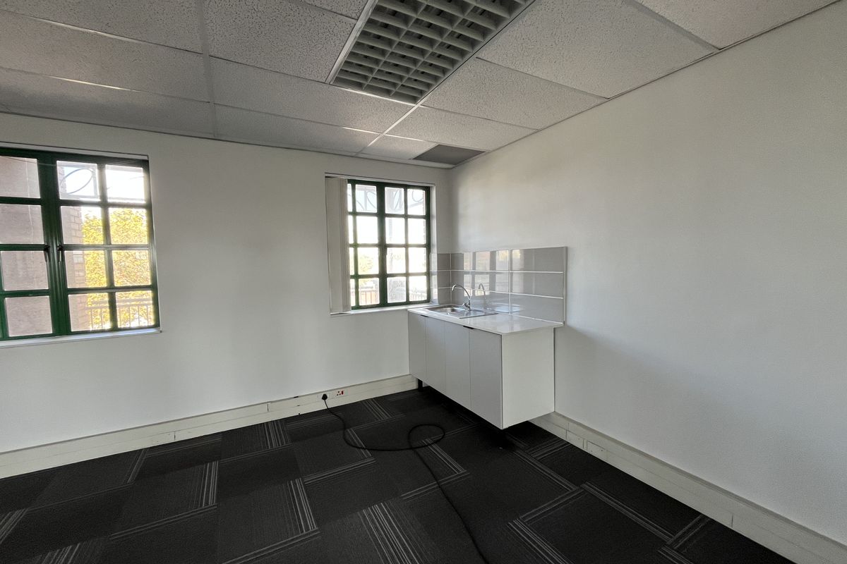 A functional office kitchenette area featuring a compact sink unit, tiled splashback, and large windows providing natural light.