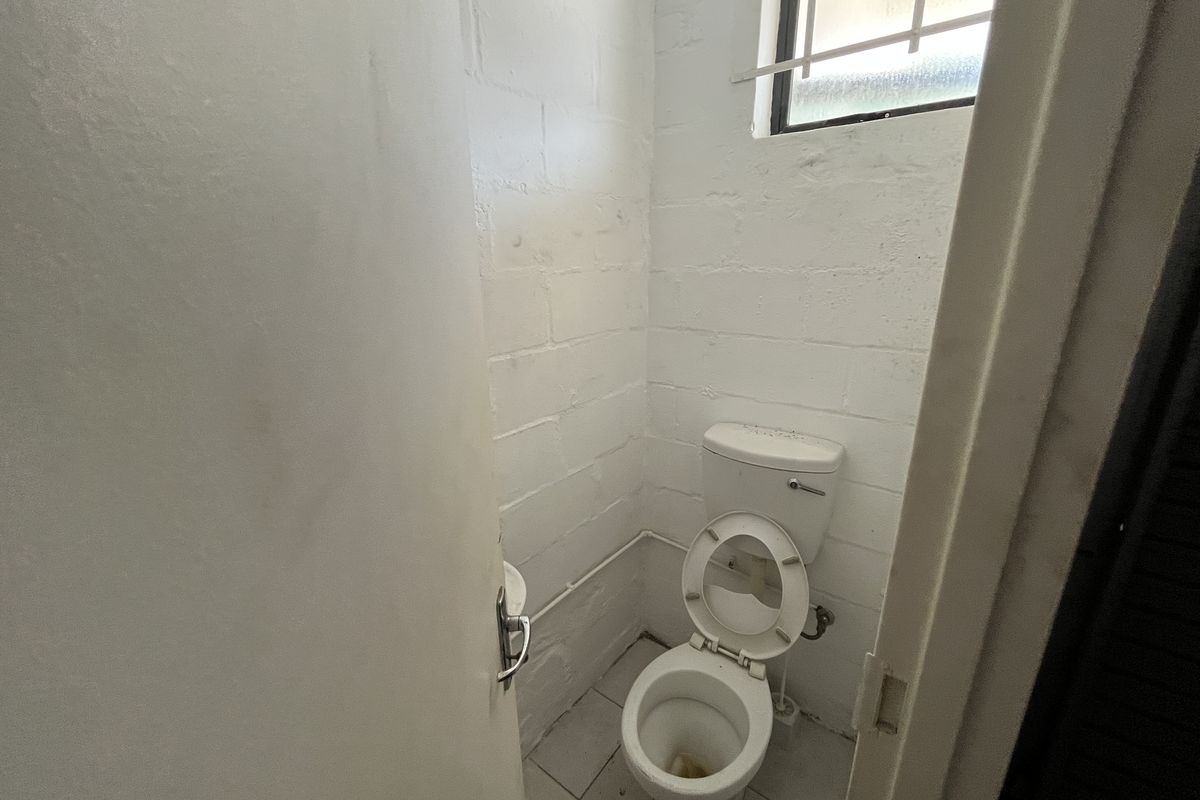 A small, functional industrial-style restroom featuring a white toilet, tiled flooring, and a high-set window with security bars.