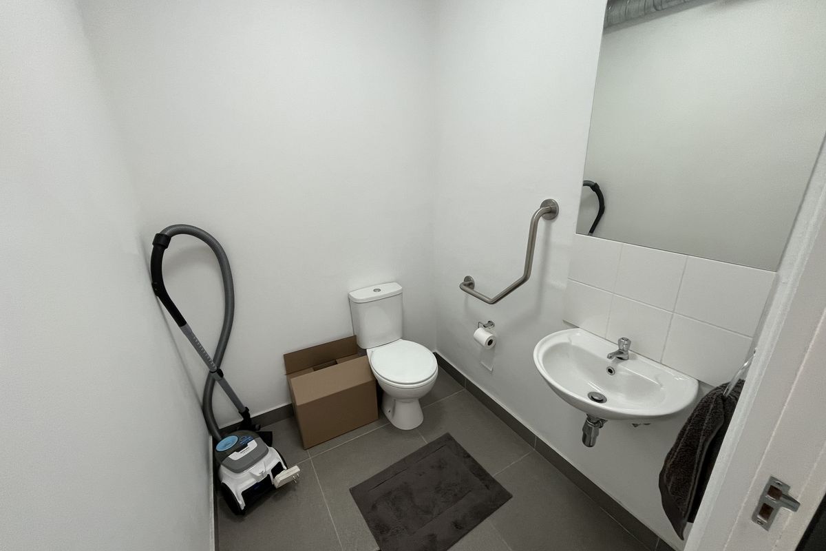 A functional accessible restroom featuring a wall-mounted basin, toilet, and safety grab rail, finished with neutral floor tiling and white walls.