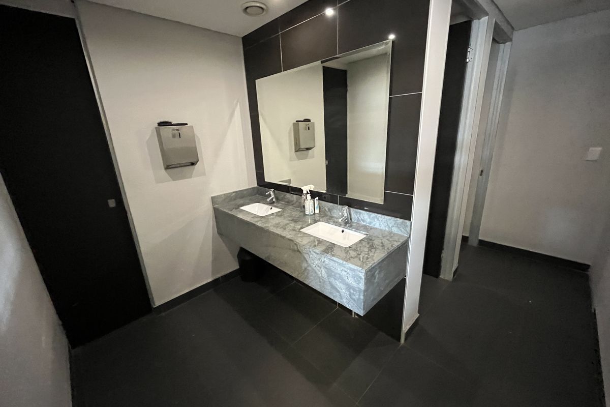 A modern office restroom featuring a dual-basin vanity with stone countertop, large wall-mounted mirror, and dark tiled flooring.