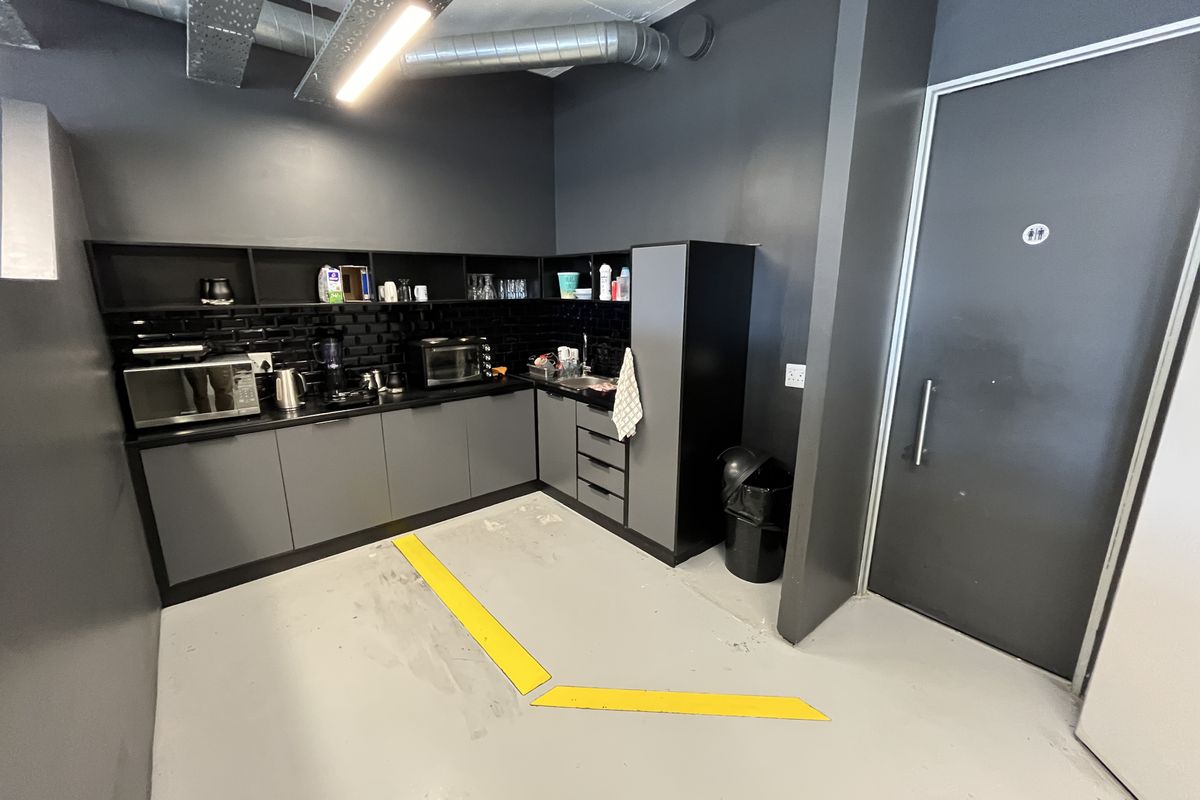 A compact office kitchenette featuring dark cabinetry, subway tile backsplash, integrated sink, and exposed industrial ceiling ducting.