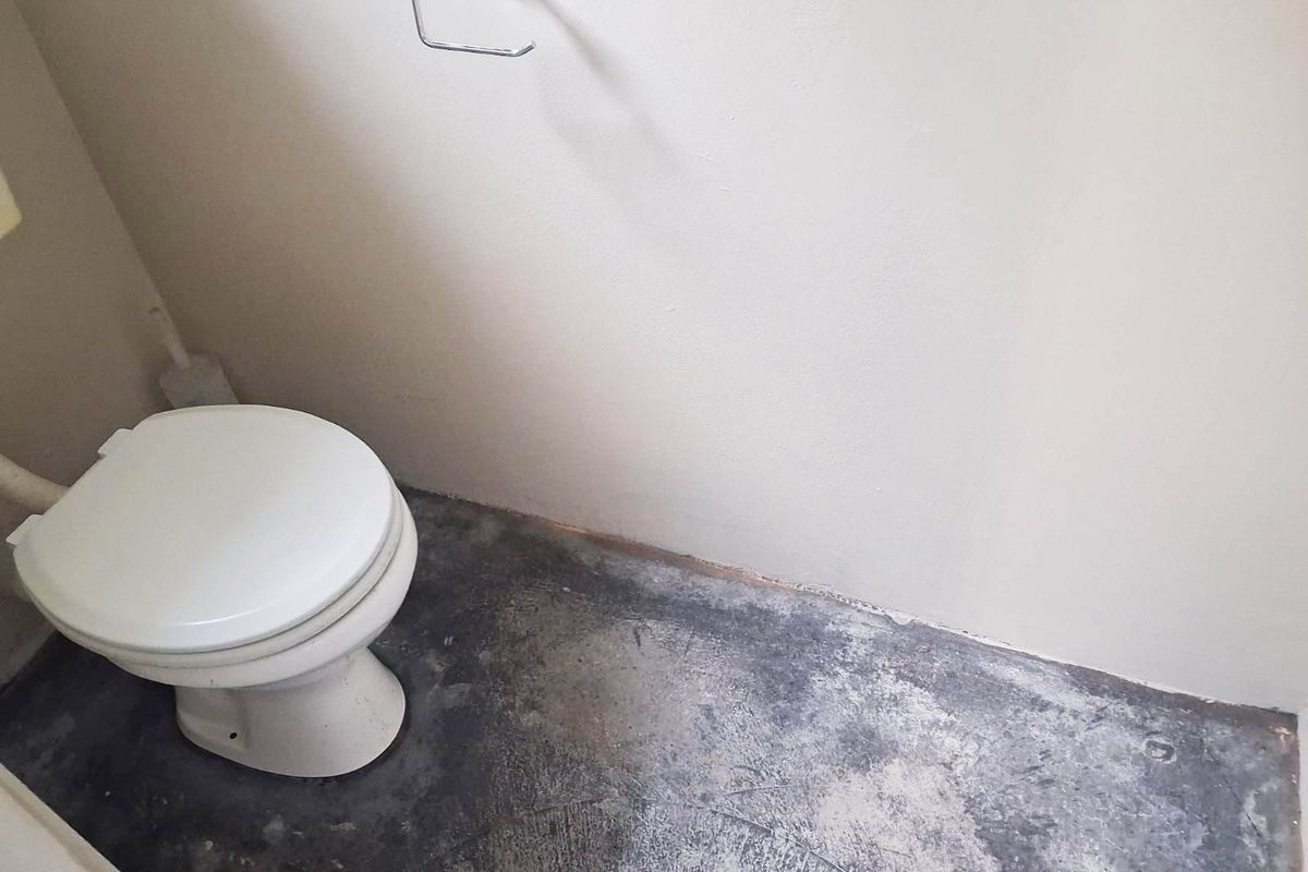 A small restroom facility featuring a standard toilet and wall-mounted toilet paper holder, currently requiring flooring and wall finish improvements.