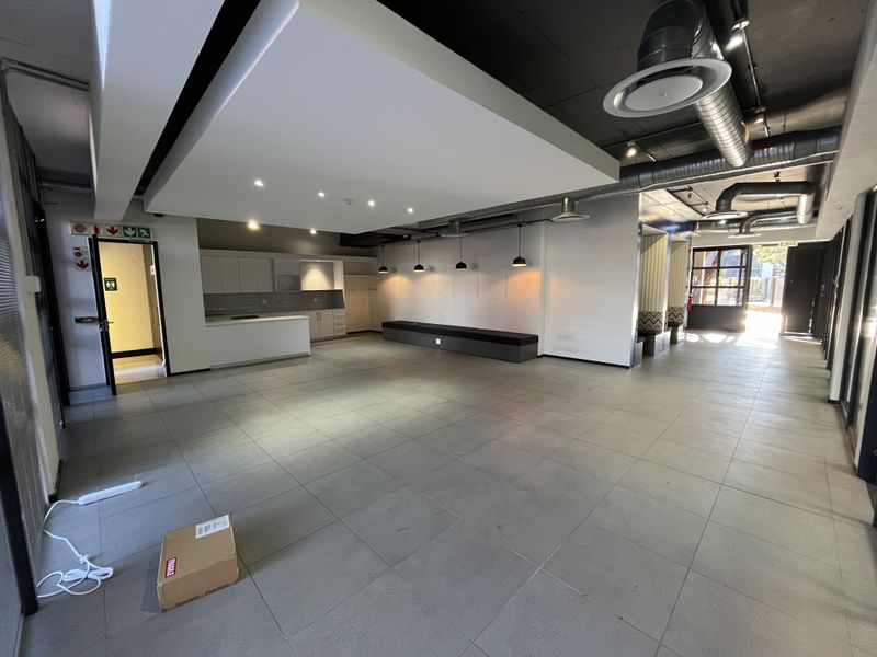 Spacious open-plan office area featuring modern tiled flooring, exposed industrial ducting, a built-in kitchenette, and integrated bench seating under a suspended ceiling feature.