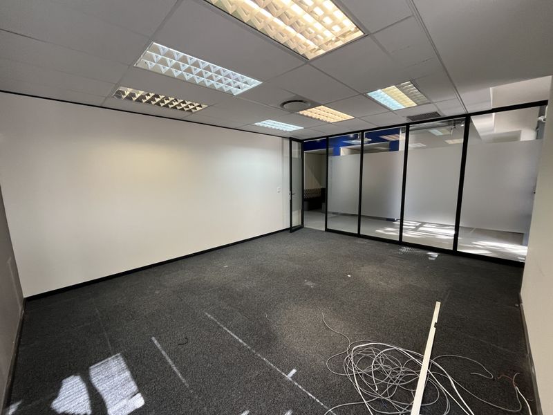 An empty office space featuring glass partition walls, suspended ceilings with recessed lighting, and dark carpeting, suitable for a professional workspace.