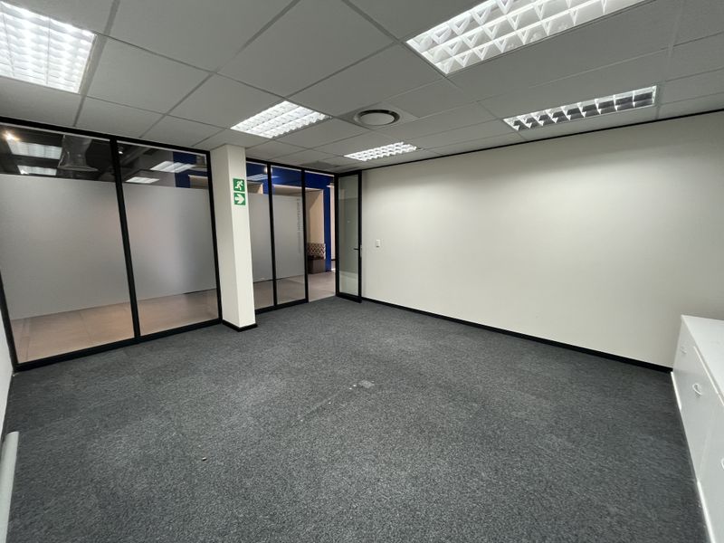 A vacant office space featuring glass partition walls, suspended ceilings with recessed lighting, and grey carpet flooring.