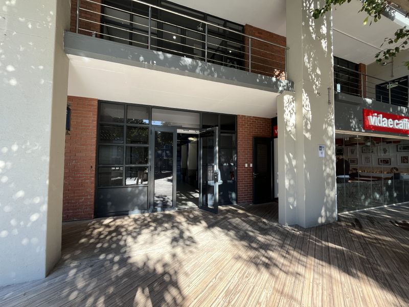 Modern office entrance featuring exposed brickwork, timber decking, and a steel-framed glass facade with an overhead balcony.
