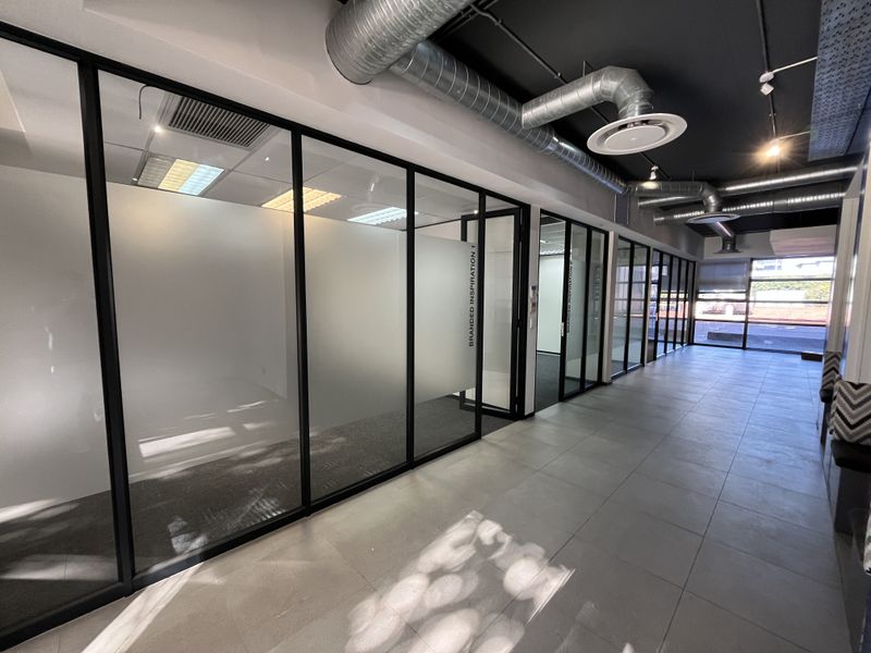 Modern office corridor featuring glass-partitioned private offices, exposed industrial-style ductwork, and a combination of tiled and carpeted flooring.