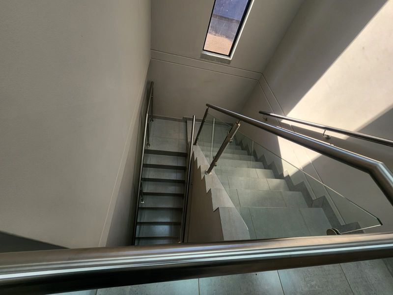 A modern industrial staircase featuring tiled steps, a sleek glass balustrade with stainless steel handrails, and a high-positioned skylight for natural illumination.