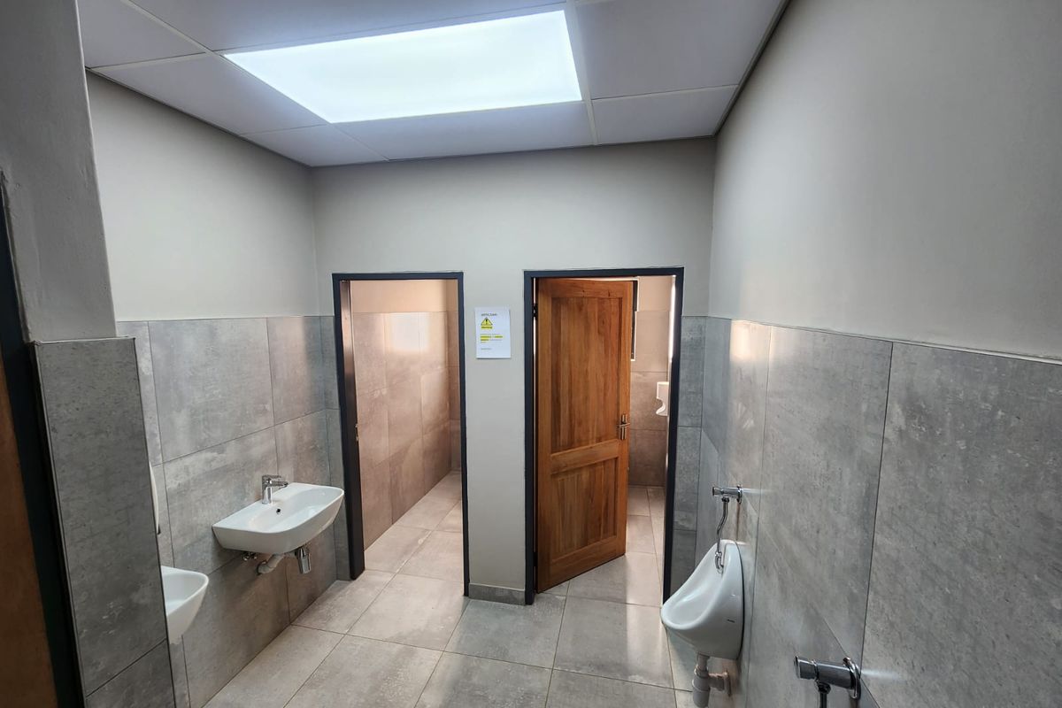 A clean, modern industrial-style communal bathroom featuring tiled walls, wall-mounted basins, urinals, and a suspended ceiling with integrated lighting.