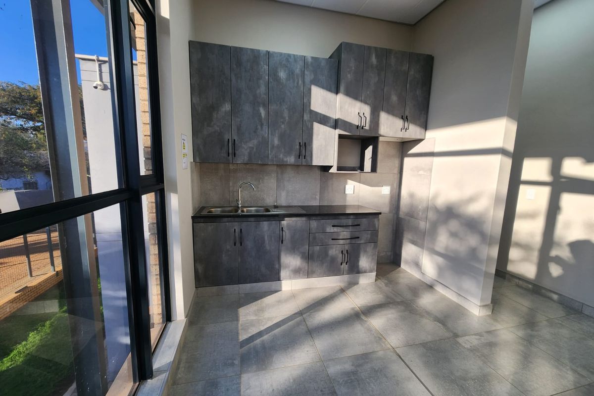 A modern, minimalist kitchenette featuring dark textured cabinetry, a stainless steel sink, and large windows providing ample natural light in an industrial setting.