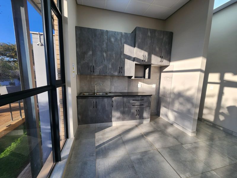 A modern, minimalist kitchenette featuring dark textured cabinetry, a stainless steel sink, and large windows providing ample natural light in an industrial setting.