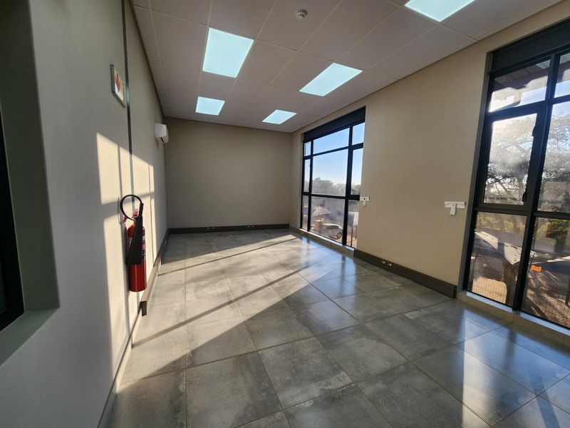 A bright, modern, and empty office space featuring large windows, tiled flooring, recessed ceiling lights, and a wall-mounted air conditioning unit.