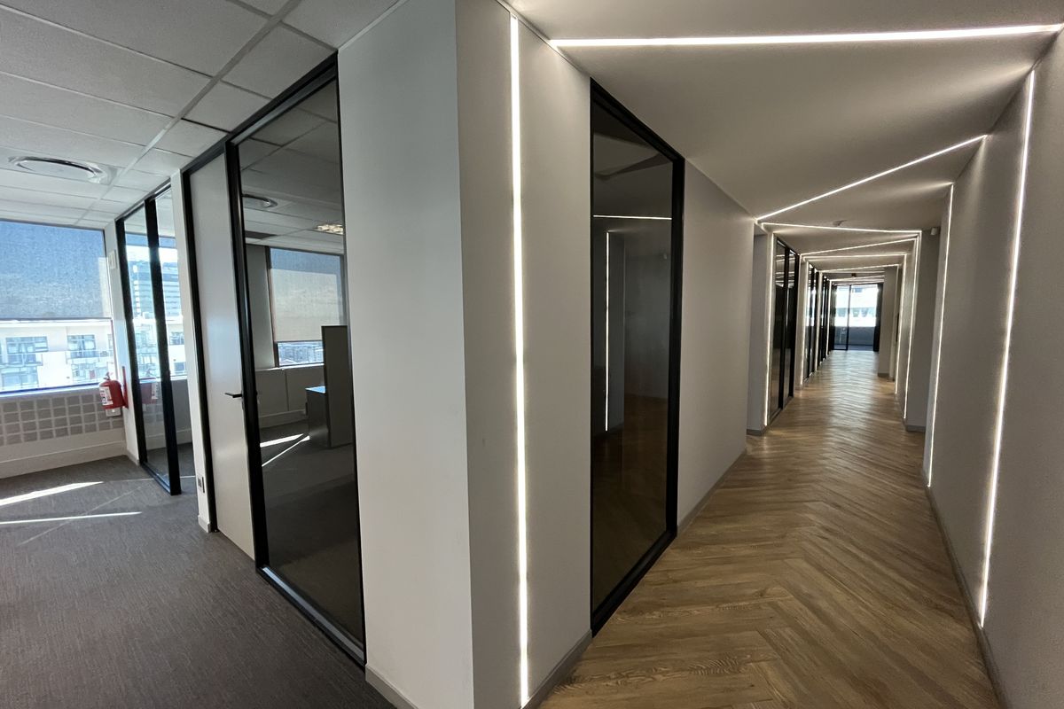 Modern office hallway featuring herringbone wood flooring, sleek glass partitions, and integrated recessed LED strip lighting for a contemporary professional aesthetic.