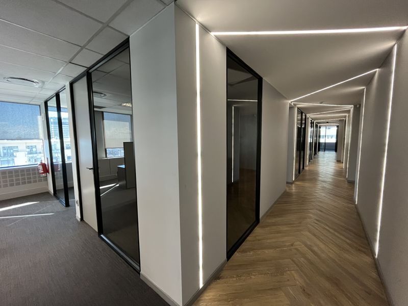 Modern office hallway featuring herringbone wood flooring, sleek glass partitions, and integrated recessed LED strip lighting for a contemporary professional aesthetic.
