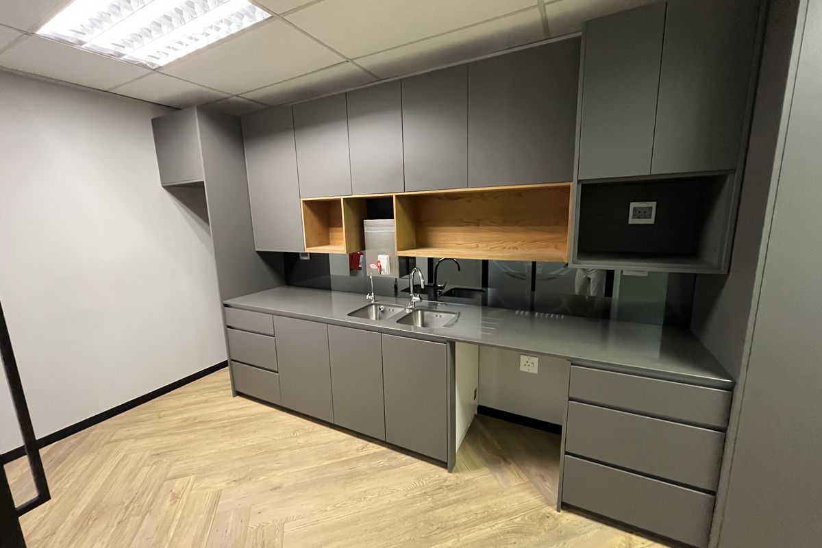 Modern office kitchenette featuring sleek grey cabinetry, wood-accented open shelving, a stainless steel sink, and durable herringbone-patterned flooring.