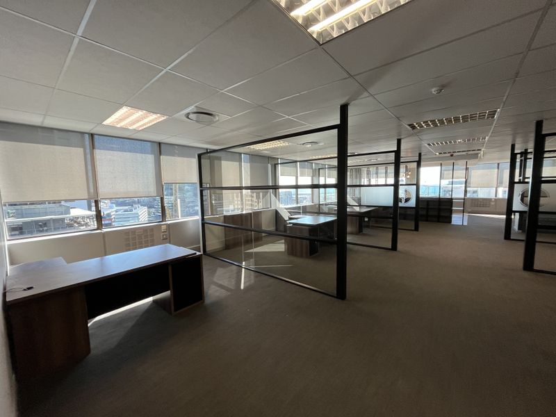 Open-plan office space featuring glass partitions, suspended ceilings with recessed lighting, carpeted flooring, and large windows with roller blinds offering city views.