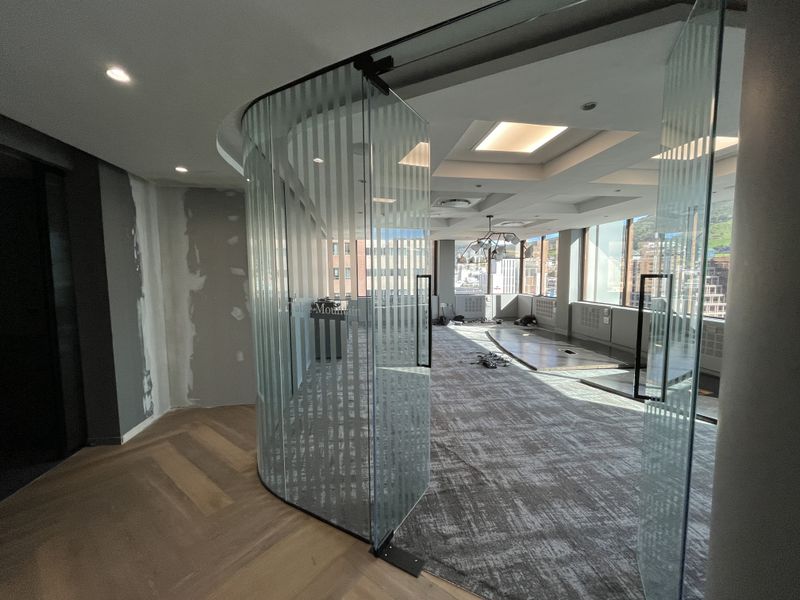 Modern office space featuring a curved glass partition, floor-to-ceiling windows with city views, and a mix of carpet and hardwood flooring.