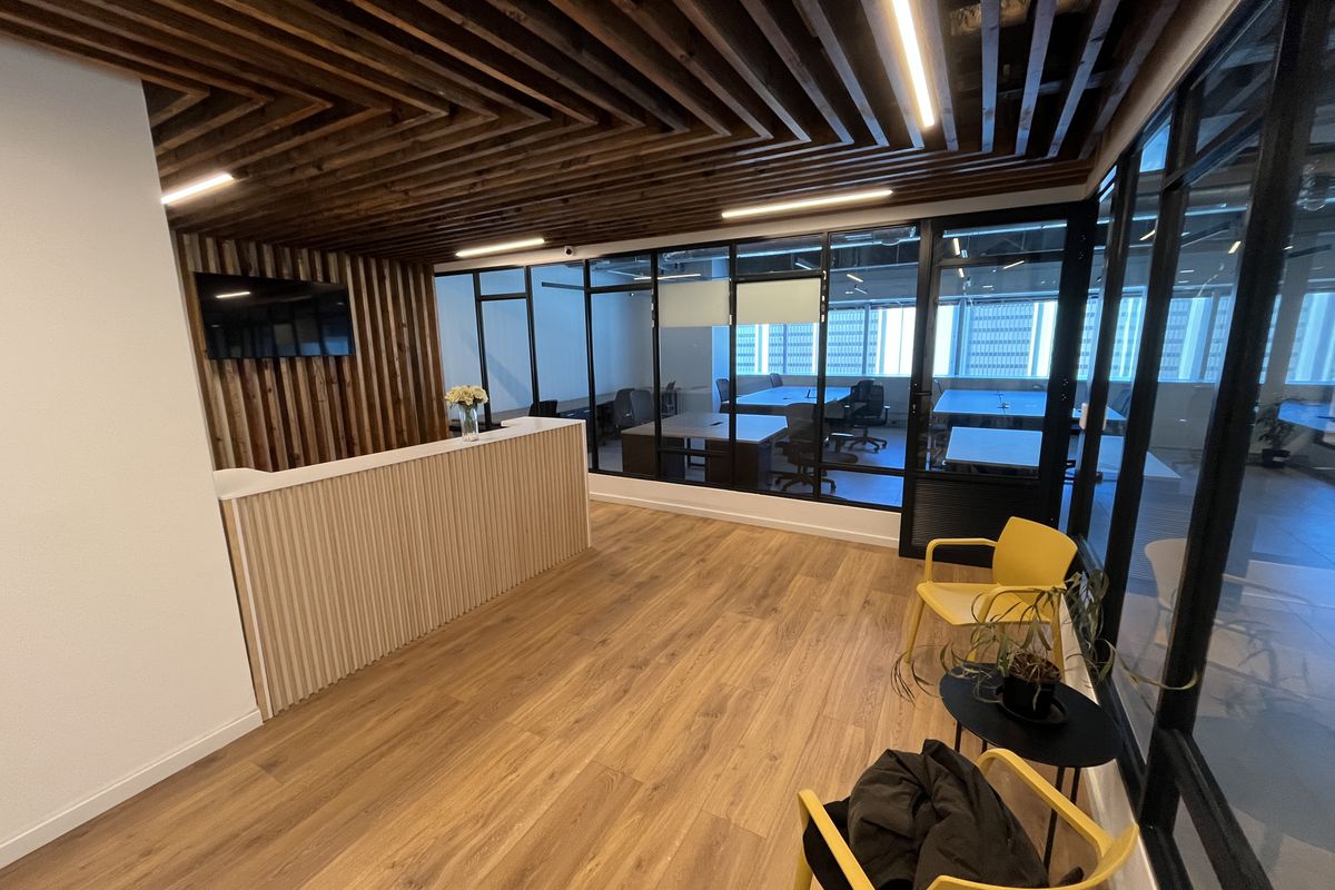 Modern office reception area featuring a custom reception desk, slatted timber ceiling, and floor-to-ceiling black framed glass partitions overlooking the workspace.