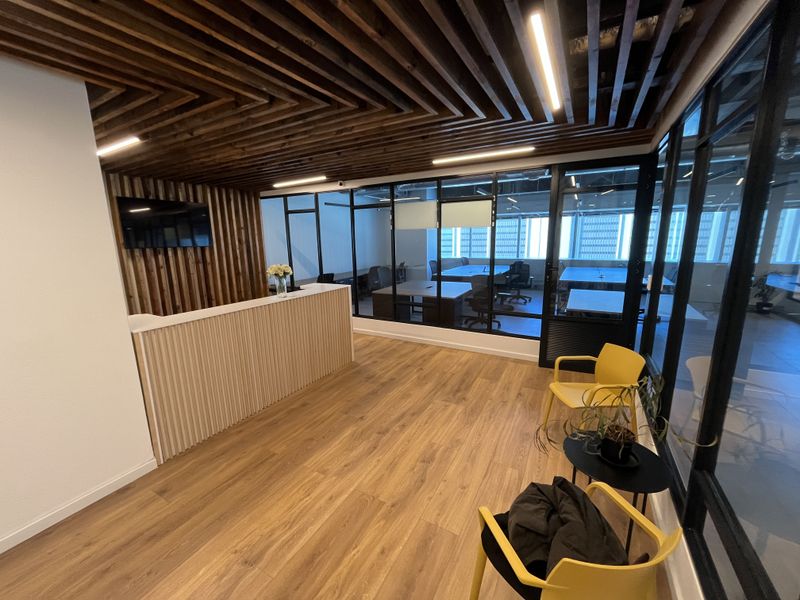 Modern office reception area featuring a custom reception desk, slatted timber ceiling, and floor-to-ceiling black framed glass partitions overlooking the workspace.
