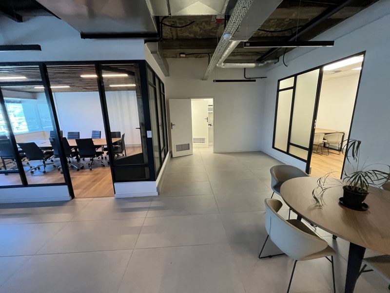 Modern office space featuring exposed industrial ceiling ducting, glass-partitioned meeting rooms, and clean tiled flooring throughout the open-plan layout.
