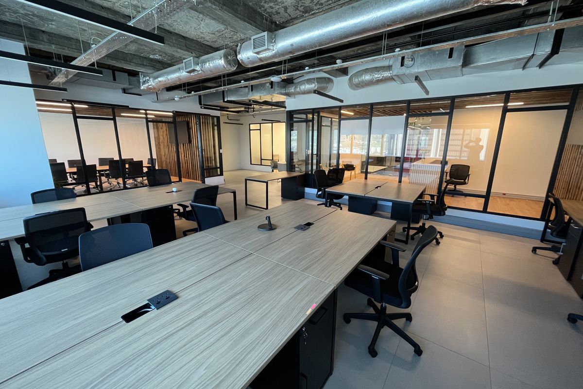 Modern open-plan office space featuring exposed industrial ceilings, glass-partitioned meeting rooms, and contemporary tiled flooring with integrated workstation power connectivity.