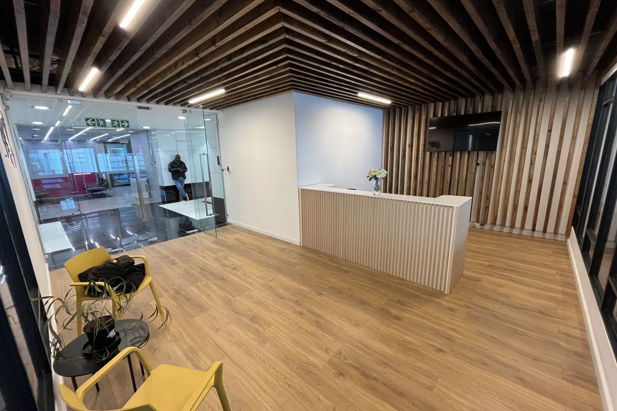 Modern office reception area featuring a custom reception desk, slatted timber ceiling, and warm wood-look flooring with glass entry doors.