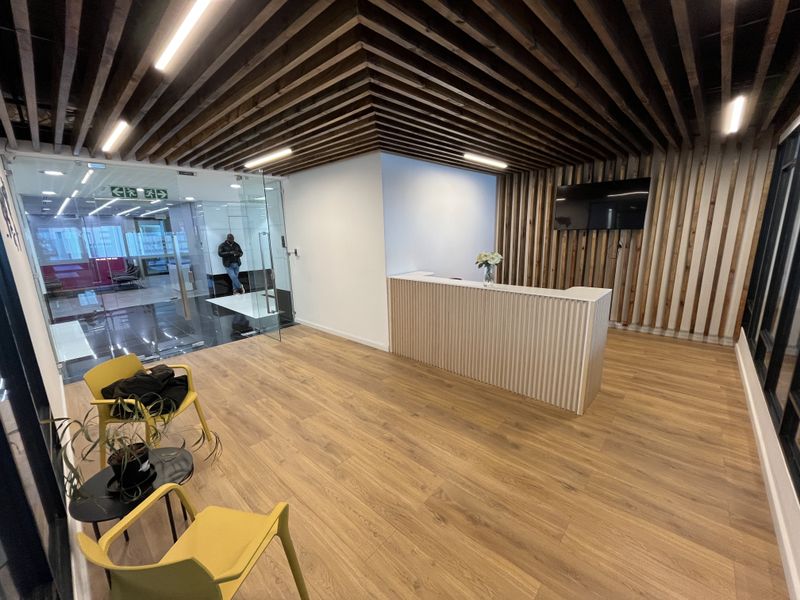 Modern office reception area featuring a custom reception desk, slatted timber ceiling, and warm wood-look flooring with glass entry doors.