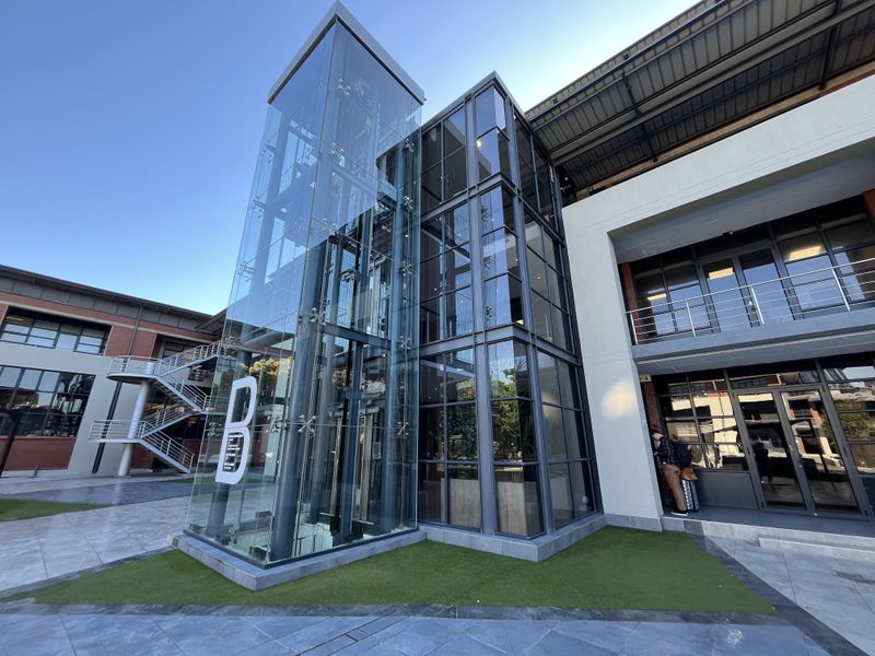 Modern office building exterior featuring a prominent glass elevator shaft, steel staircase, and contemporary curtain wall glazing in a landscaped courtyard setting.