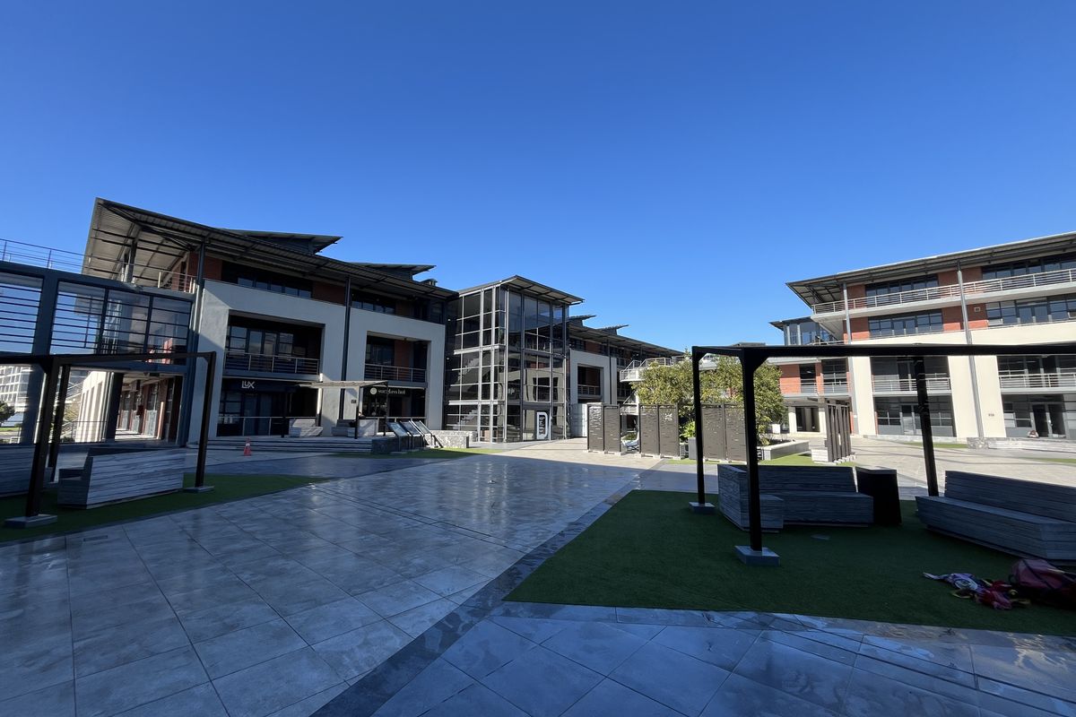 A modern office complex courtyard featuring tiled paving, artificial turf areas, pergola structures, and multi-story buildings with glass facades and balconies.