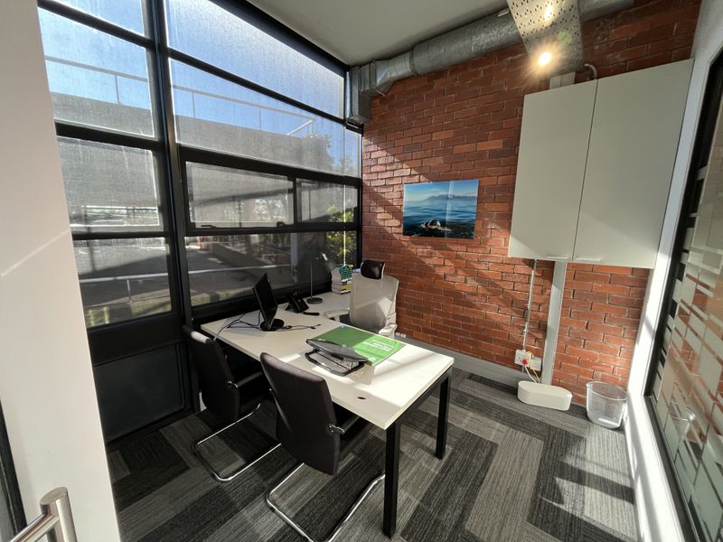 A modern office space featuring an exposed brick wall, large windows providing natural light, exposed industrial ceiling ducting, and professional carpet tiles.
