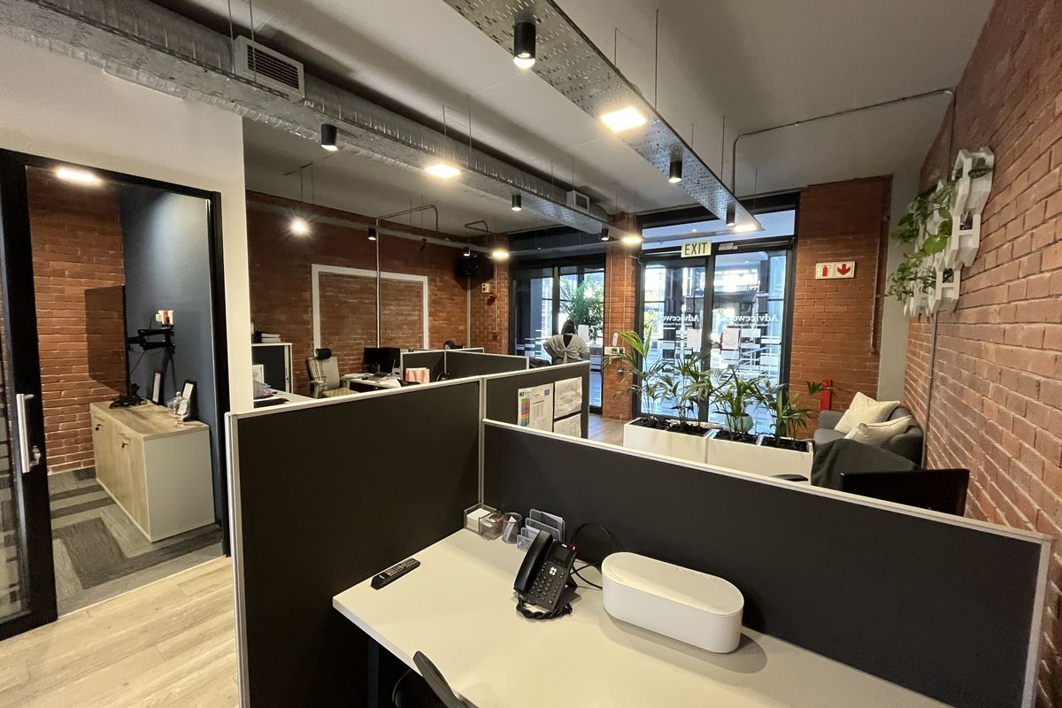 Modern open-plan office space featuring exposed brick walls, industrial-style ceiling ducting, integrated lighting, and partitioned workstations with laminate flooring.