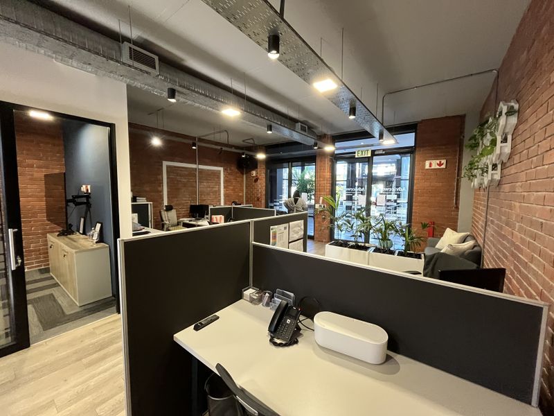 Modern open-plan office space featuring exposed brick walls, industrial-style ceiling ducting, integrated lighting, and partitioned workstations with laminate flooring.