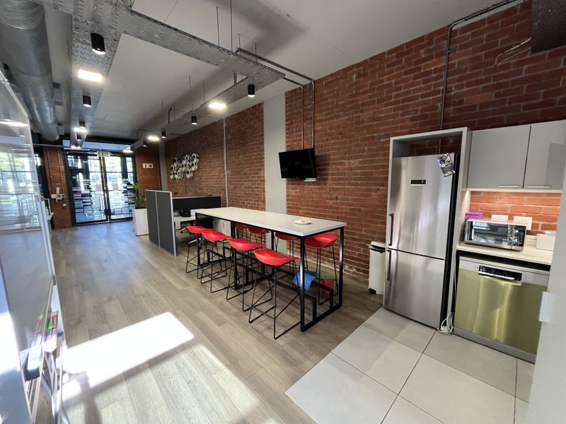 Modern open-plan office space featuring exposed brick walls, industrial ceiling ducting, and a compact kitchenette with integrated appliances.