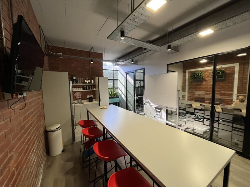 Modern office breakout area featuring exposed brick walls, industrial-style ceiling ducting, a glass-partitioned meeting room, and a compact kitchenette.