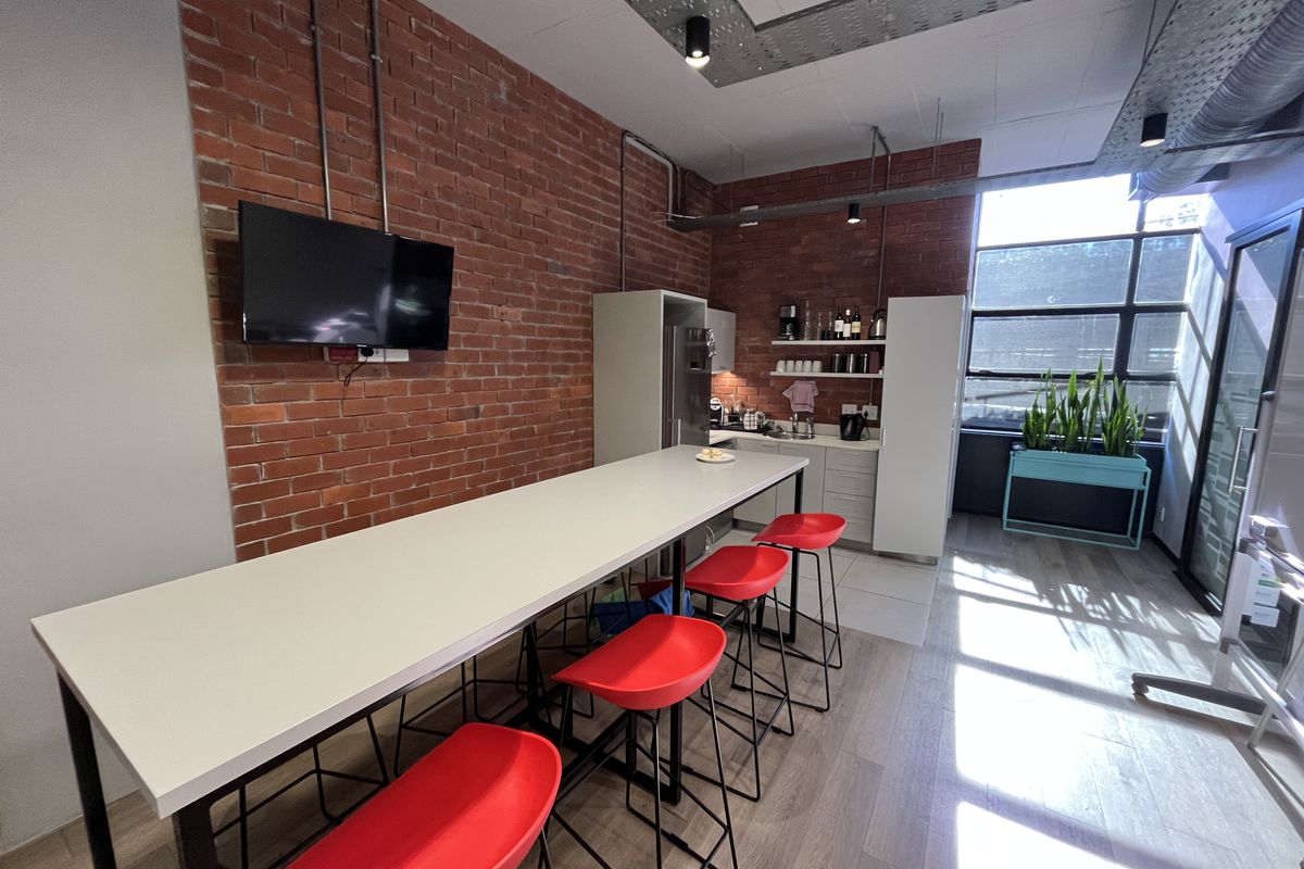 Modern office kitchenette featuring exposed brick walls, industrial ceiling ducting, built-in cabinetry, and a large window providing ample natural light.