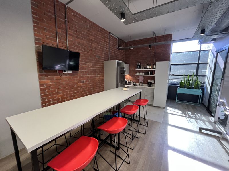 Modern office kitchenette featuring exposed brick walls, industrial ceiling ducting, built-in cabinetry, and a large window providing ample natural light.
