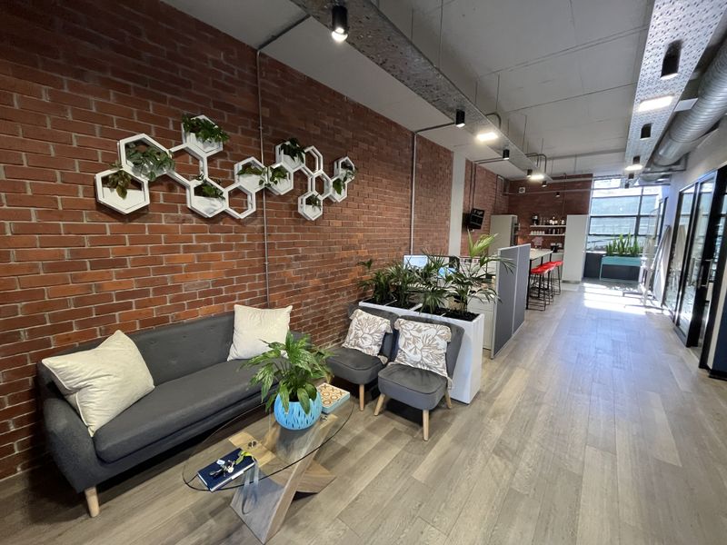 Modern open-plan office space featuring exposed brick walls, industrial-style ceiling ducting, track lighting, and a functional kitchenette area with sleek flooring.