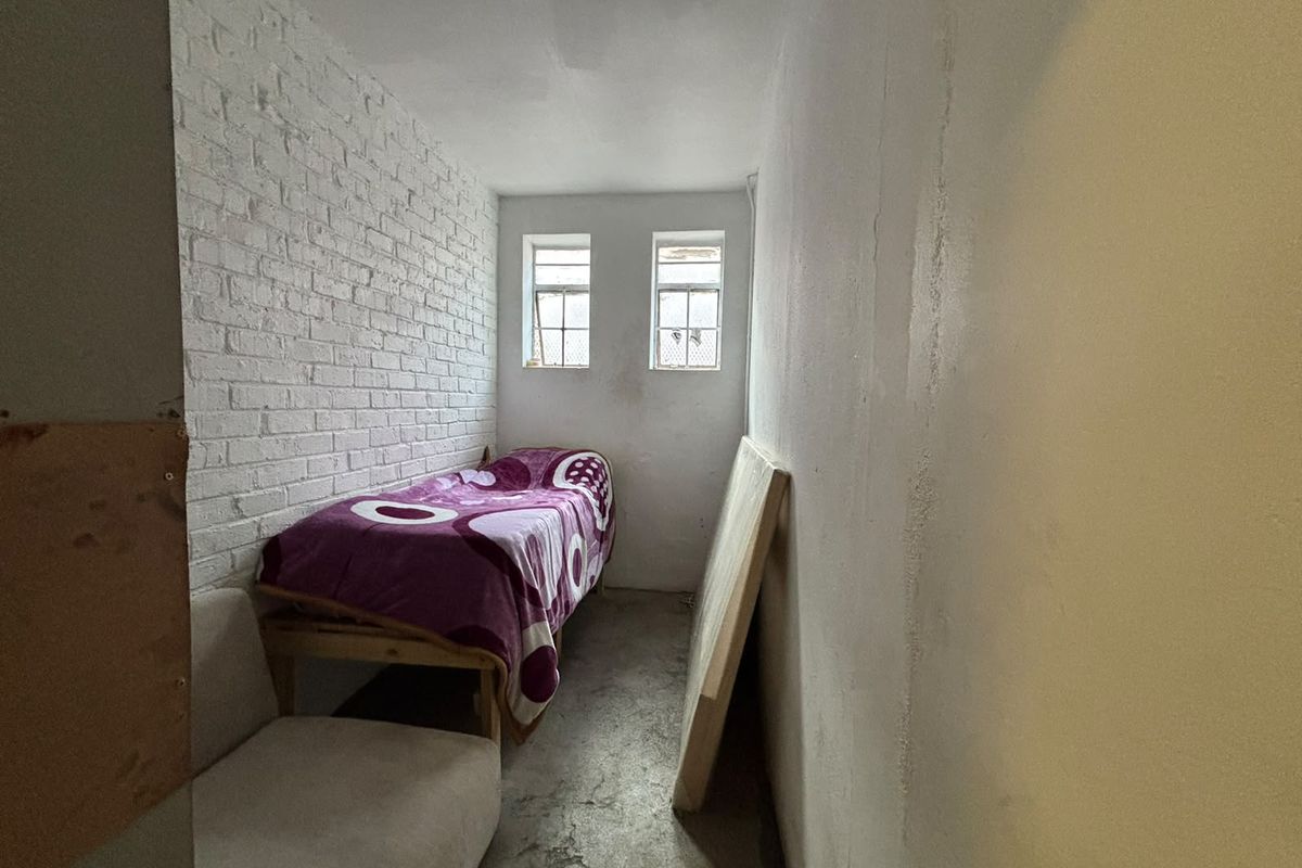 A small, industrial-style bedroom featuring an exposed white brick wall, concrete flooring, and two small casement windows.