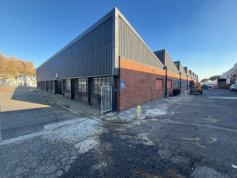 Exterior view of an industrial warehouse building featuring a sawtooth roof, brick and corrugated metal facade, and surrounding asphalt yard area.
