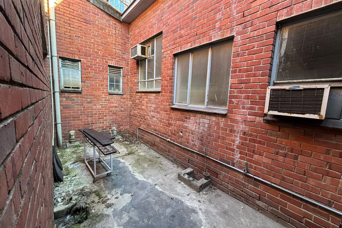 A narrow, enclosed rear exterior area featuring exposed brick walls, concrete flooring, and multiple window-mounted air conditioning units in need of maintenance.
