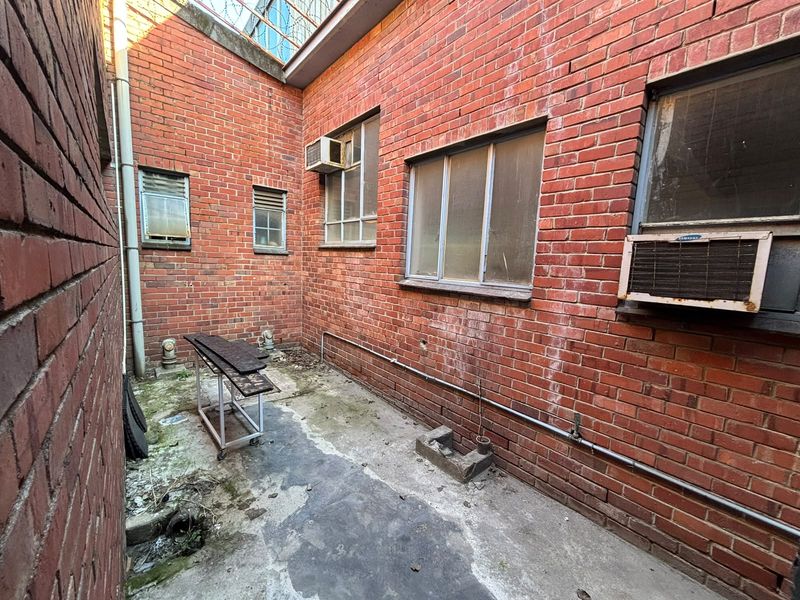 A narrow, enclosed rear exterior area featuring exposed brick walls, concrete flooring, and multiple window-mounted air conditioning units in need of maintenance.