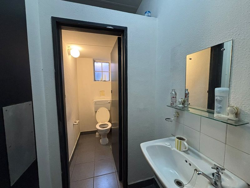 A functional bathroom featuring a wall-mounted basin, mirror, and a separate toilet area with tiled flooring and basic lighting.