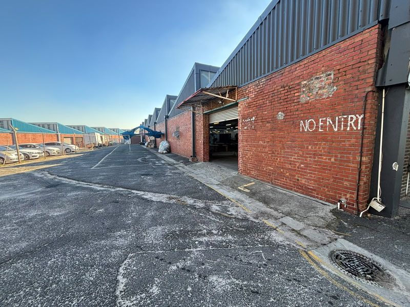 Exterior view of an industrial warehouse facility featuring red brick construction, metal roofing, and a roller shutter door access point.