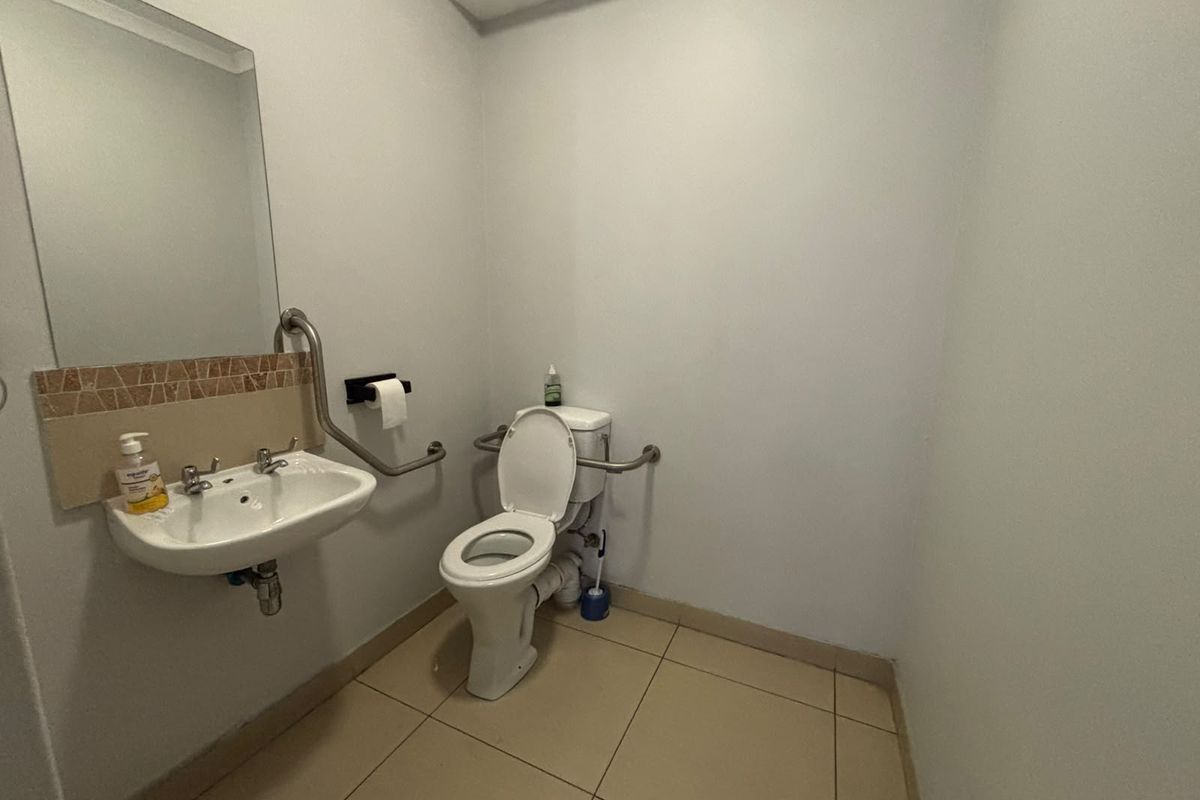 A functional, accessible bathroom featuring a wall-mounted basin, toilet, and safety grab rails with neutral tiled flooring and splashback.
