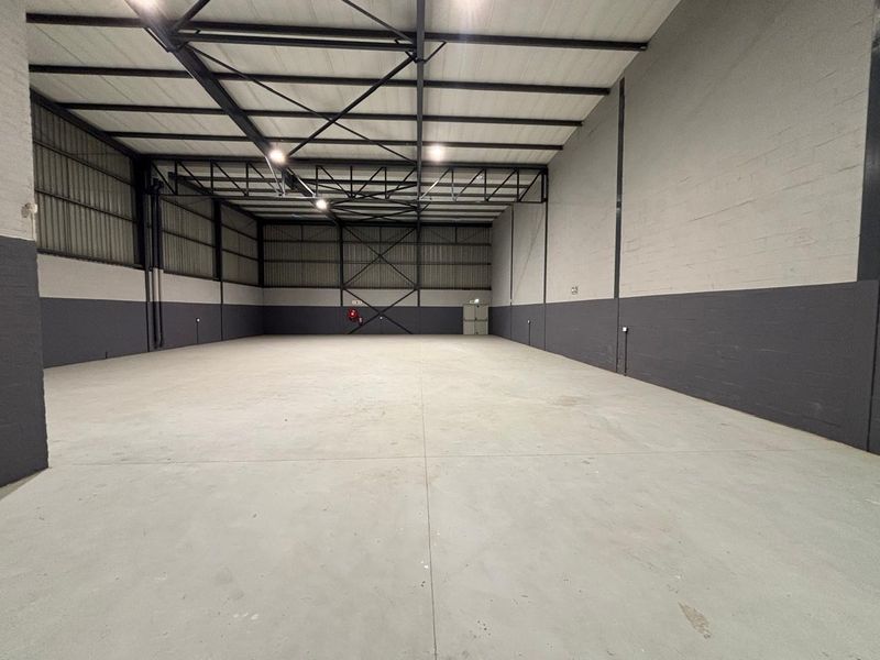 A spacious, clean industrial warehouse unit featuring high ceilings with exposed steel trusses, concrete flooring, and freshly painted masonry walls.
