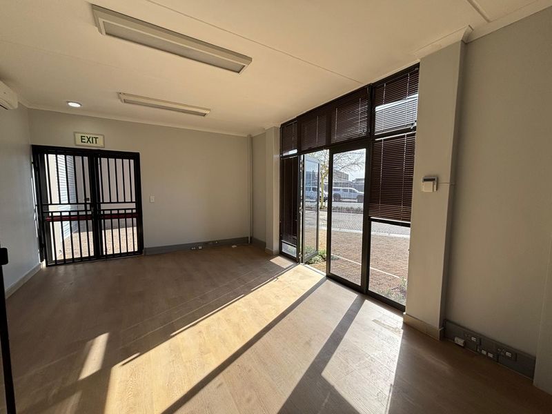 A bright, empty office space featuring laminate flooring, large windows with blinds, security gate, and integrated electrical trunking.