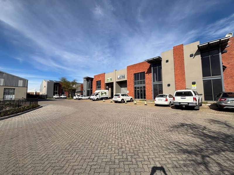A modern industrial complex featuring a paved driveway, brick and concrete facade, and multiple roller shutter access points for commercial or warehouse use.