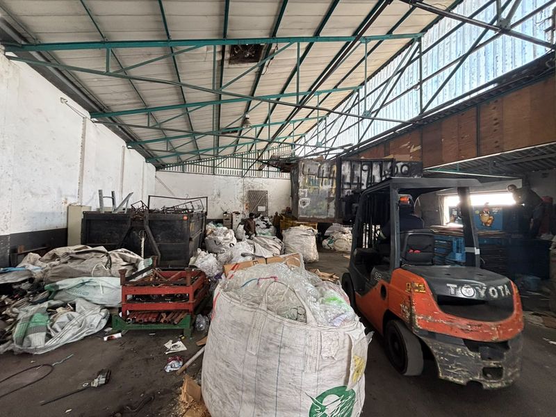 Large industrial warehouse space featuring high exposed steel roof trusses, corrugated metal roofing, and concrete flooring, currently utilized for storage and material handling.