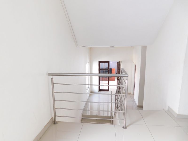A bright, minimalist hallway featuring tiled flooring and a modern stainless steel balustrade leading to a glass-panelled doorway.
