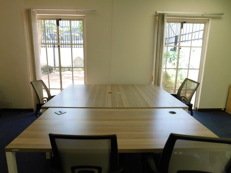 A functional office space featuring dual workstations, carpeted flooring, and large windows with security bars overlooking an outdoor area.